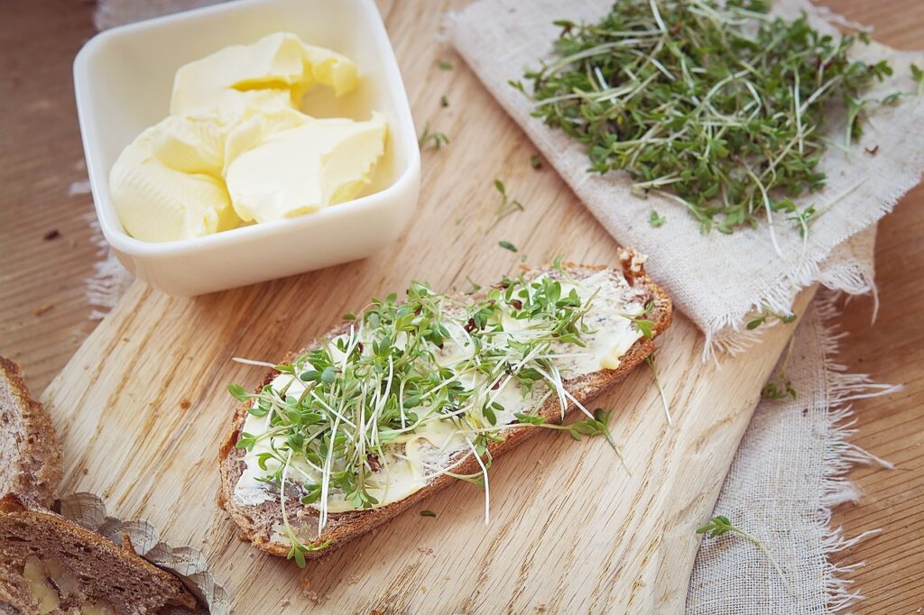 A slice of whole-grain bread spread with butter and topped with fresh cress, placed on a wooden cutting board next to a bowl of butter and a pile of cress.