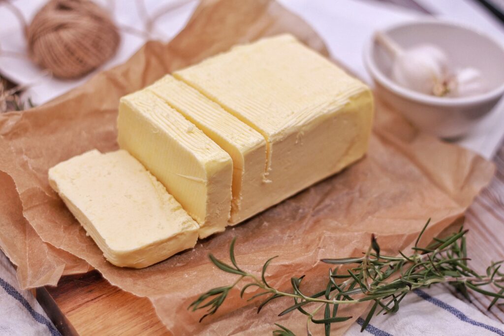 A block of butter sliced on a piece of parchment paper, surrounded by fresh rosemary sprigs, with a garlic bulb in a small bowl in the background.